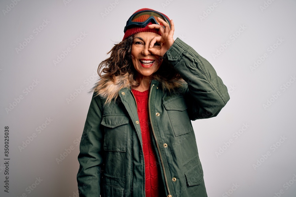 Middle age skier woman wearing snow sportswear and ski goggles over white background doing ok gesture with hand smiling, eye looking through fingers with happy face. Middle age skier woman wearing snow sportswear and ski goggles over white background doing ok gesture with hand smiling, eye looking through fingers with happy face.