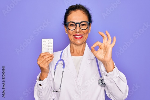 Middle age senior professional doctor woman holding pharmaceutical pills doing ok sign with fingers  excellent symbol
