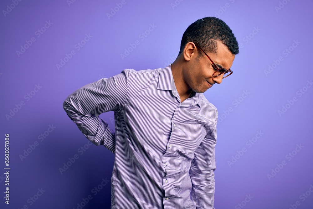 Handsome african american man wearing striped shirt and glasses over purple background Suffering of backache, touching back with hand, muscular pain Handsome african american man wearing striped shirt and glasses over purple background Suffering of backache, touching back with hand, muscular pain