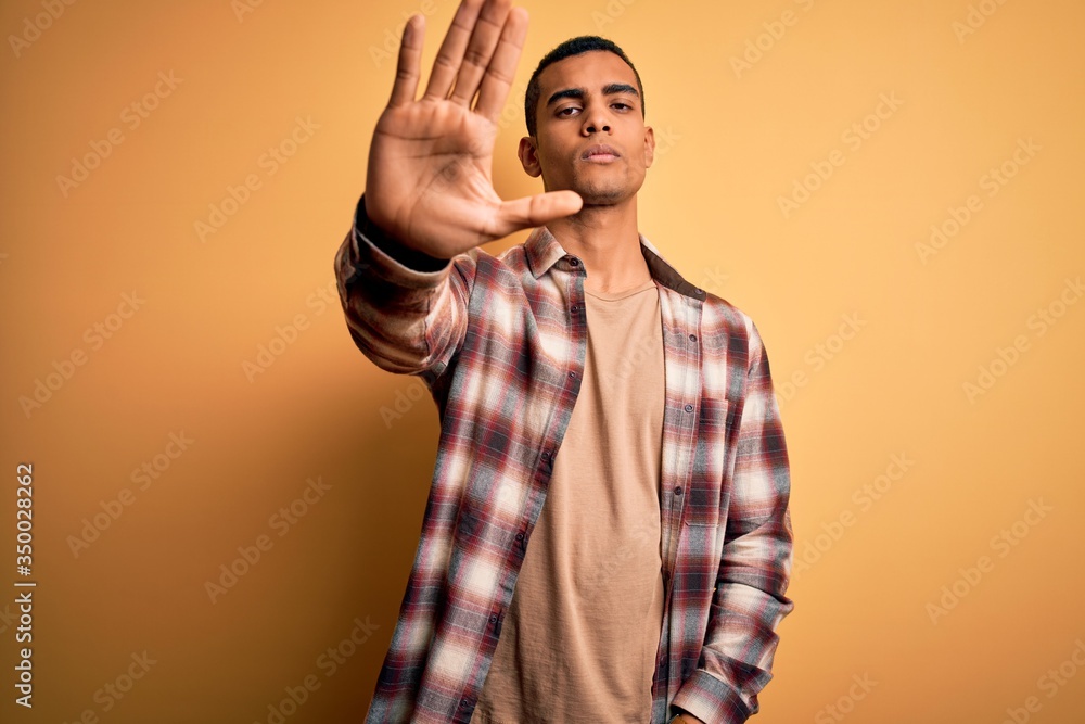Young handsome african american man wearing casual shirt standing over yellow background doing stop sing with palm of the hand. Warning expression with negative and serious gesture on the face.