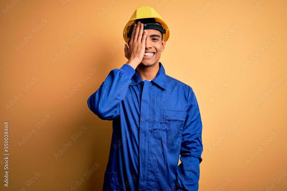 Young handsome african american worker man wearing blue uniform and security helmet covering one eye with hand, confident smile on face and surprise emotion.