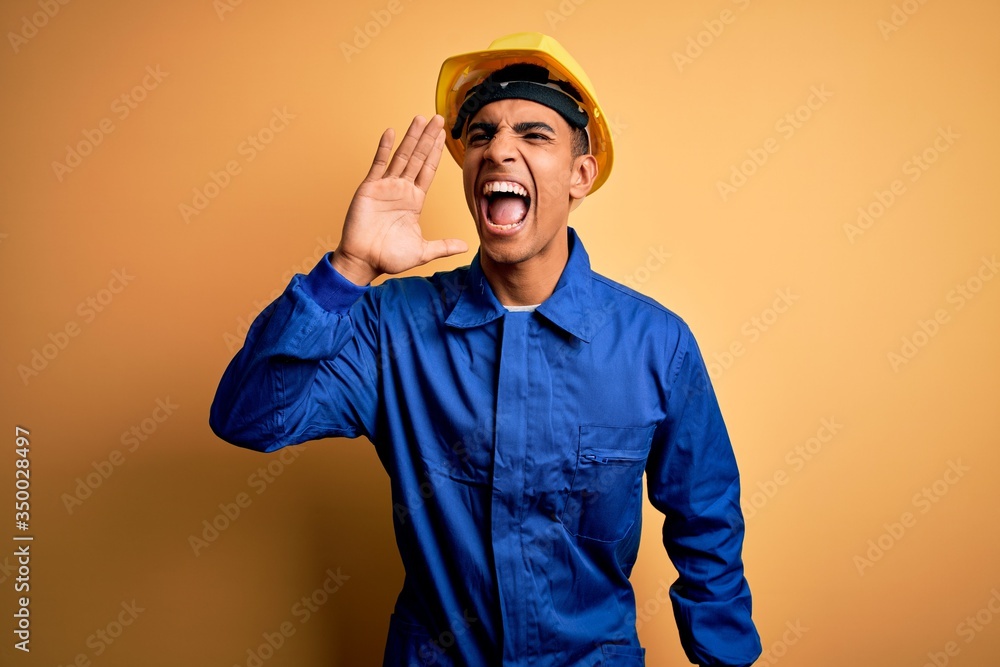 Young handsome african american worker man wearing blue uniform and security helmet shouting and screaming loud to side with hand on mouth. Communication concept.