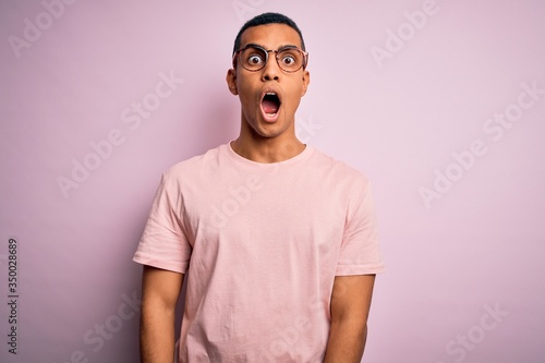 Handsome african american man wearing casual t-shirt and glasses over pink background afraid and shocked with surprise and amazed expression, fear and excited face.