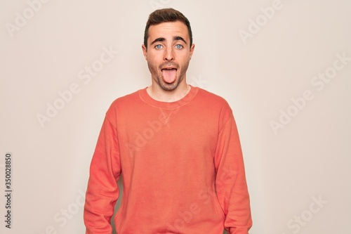 Young handsome man with blue eyes wearing casual sweater standing over white background sticking tongue out happy with funny expression. Emotion concept.