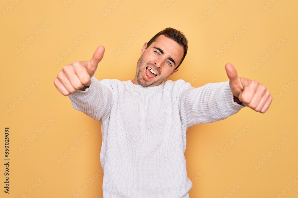 Young handsome man with blue eyes wearing casual sweater standing over yellow background approving doing positive gesture with hand, thumbs up smiling and happy for success. Winner gesture.
