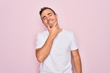 Handsome man with blue eyes wearing casual white t-shirt standing over pink background looking confident at the camera smiling with crossed arms and hand raised on chin. Thinking positive.