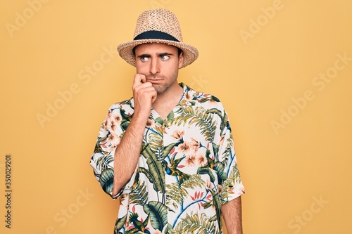 Young handsome man with blue eyes on vacation wearing summer florar shirt and hat with hand on chin thinking about question, pensive expression. Smiling with thoughtful face. Doubt concept.