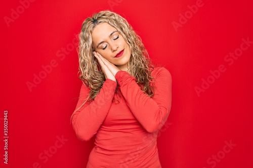 Young beautiful blonde woman wearing red casual t-shirt standing over isolated background sleeping tired dreaming and posing with hands together while smiling with closed eyes. © Krakenimages.com