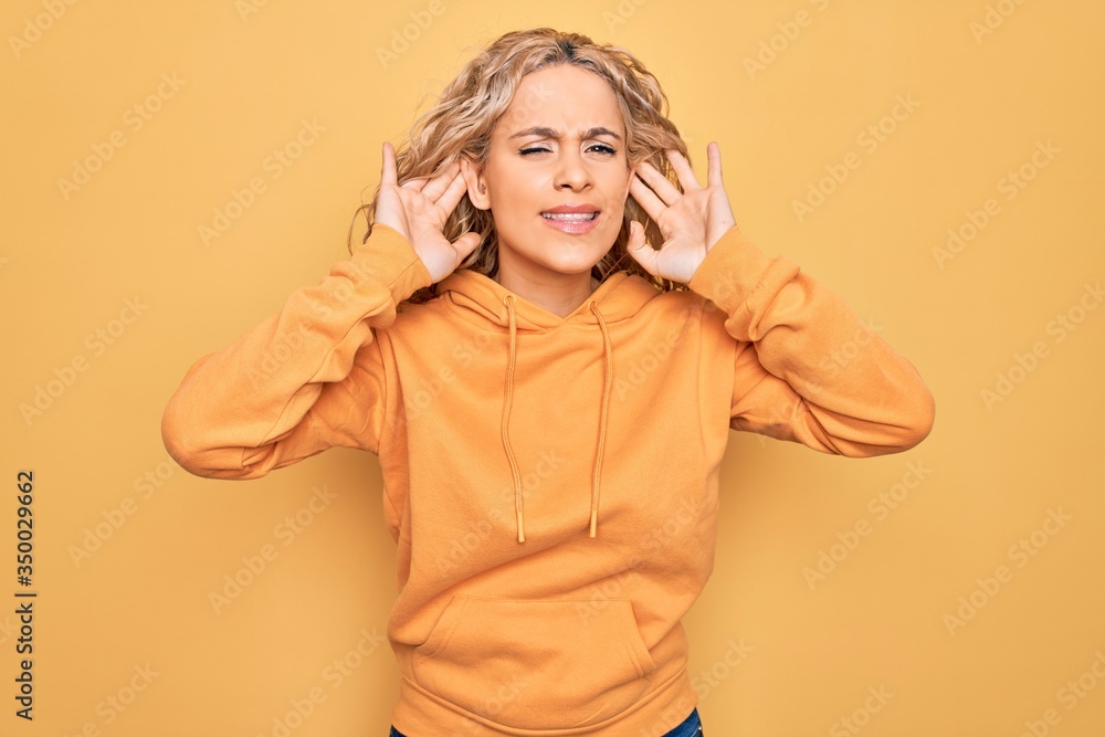 Young beautiful blonde sporty woman wearing casual sweatshirt over yellow background Trying to hear both hands on ear gesture, curious for gossip. Hearing problem, deaf Young beautiful blonde sporty woman wearing casual sweatshirt over yellow background Trying to hear both hands on ear gesture, curious for gossip. Hearing problem, deaf