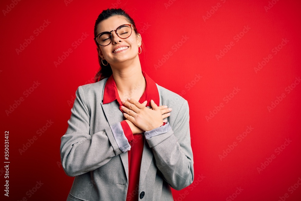 Φωτογραφία Young beautiful brunette businesswoman wearing jacket and glasses over red background smiling with hands on chest with closed eyes and grateful gesture on face