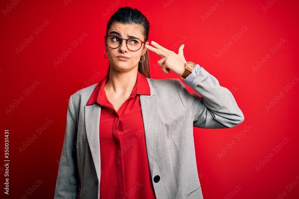 Young beautiful brunette businesswoman wearing jacket and glasses over red background Shooting and killing oneself pointing hand and fingers to head like gun, suicide gesture. Young beautiful brunette businesswoman wearing jacket and glasses over red background Shooting and killing oneself pointing hand and fingers to head like gun, suicide gesture.