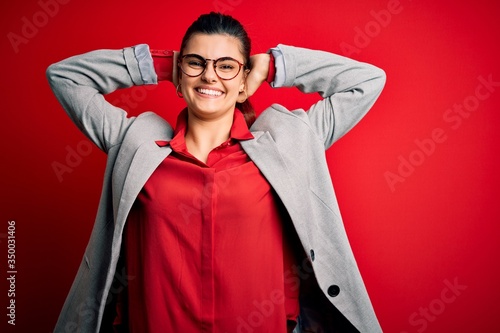 Young beautiful brunette businesswoman wearing jacket and glasses over red background relaxing and stretching, arms and hands behind head and neck smiling happy
