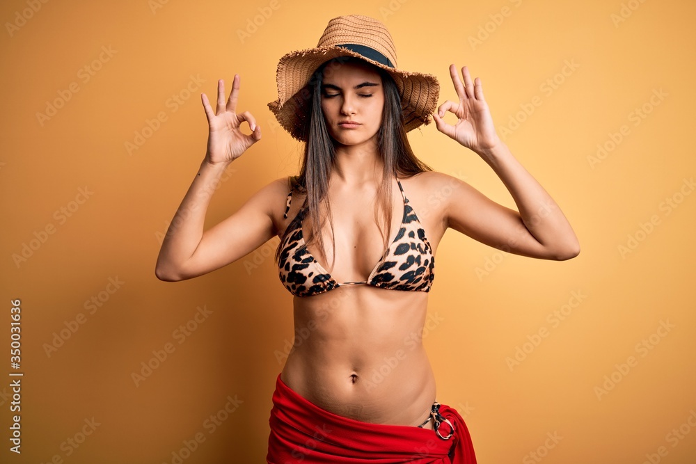 Young beautiful brunette woman on vacation wearing swimsuit and summer hat relax and smiling with eyes closed doing meditation gesture with fingers. Yoga concept. Young beautiful brunette woman on vacation wearing swimsuit and summer hat relax and smiling with eyes closed doing meditation gesture with fingers. Yoga concept.