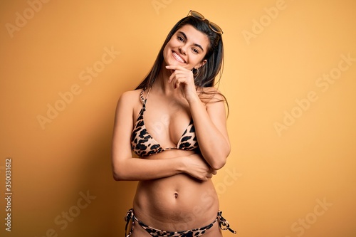 Young beautiful brunette woman on vacation wearing swimwear bikini over yellow background looking confident at the camera with smile with crossed arms and hand raised on chin. Thinking positive.