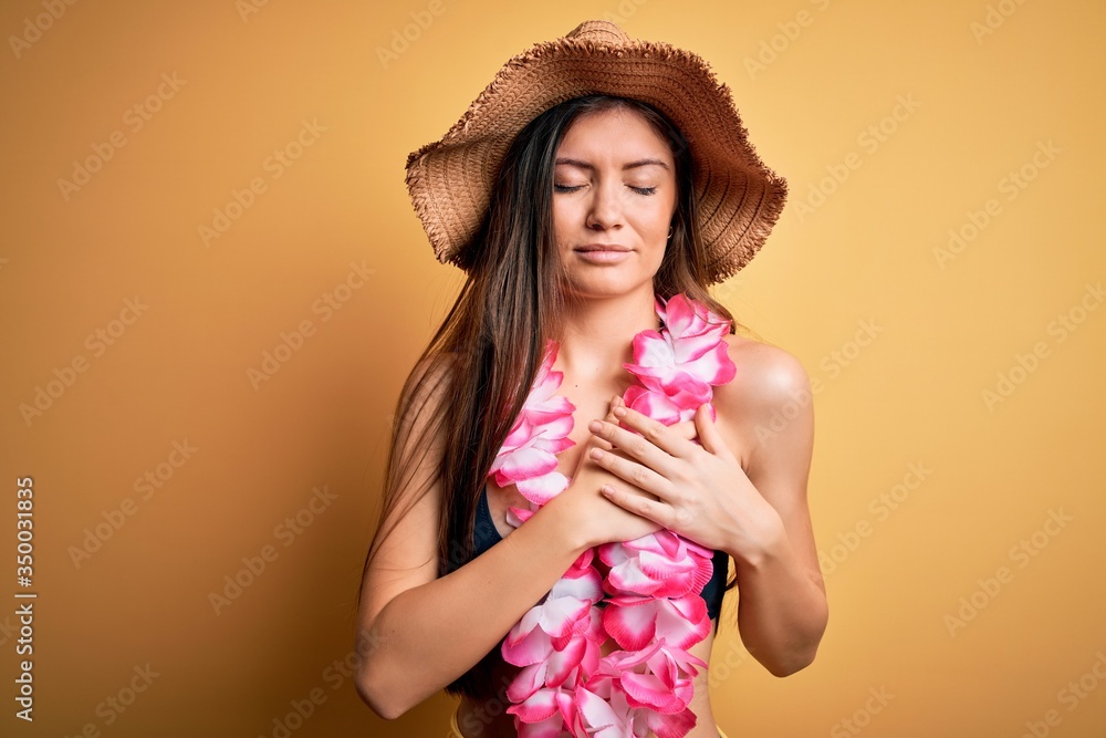 Young beautiful woman with blue eyes on vacation wearing bikini and hawaiian lei smiling with hands on chest with closed eyes and grateful gesture on face. Health concept. Young beautiful woman with blue eyes on vacation wearing bikini and hawaiian lei smiling with hands on chest with closed eyes and grateful gesture on face. Health concept.