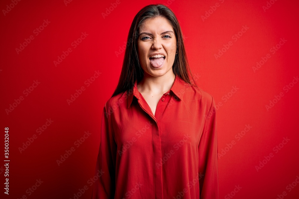 Young beautiful woman with blue eyes wearing casual shirt standing over red background sticking tongue out happy with funny expression. Emotion concept. Young beautiful woman with blue eyes wearing casual shirt standing over red background sticking tongue out happy with funny expression. Emotion concept.