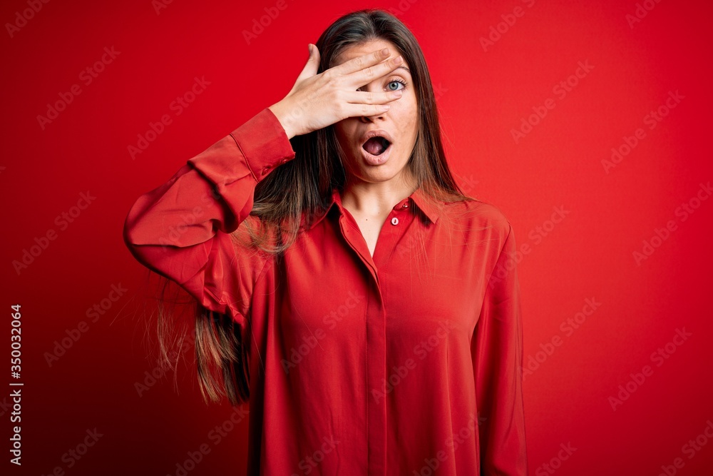 Young beautiful woman with blue eyes wearing casual shirt standing over red background peeking in shock covering face and eyes with hand, looking through fingers with embarrassed expression. Young beautiful woman with blue eyes wearing casual shirt standing over red background peeking in shock covering face and eyes with hand, looking through fingers with embarrassed expression.