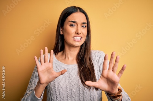 Young beautiful brunette woman wearing white casual sweater over yellow background afraid and terrified with fear expression stop gesture with hands, shouting in shock. Panic concept.