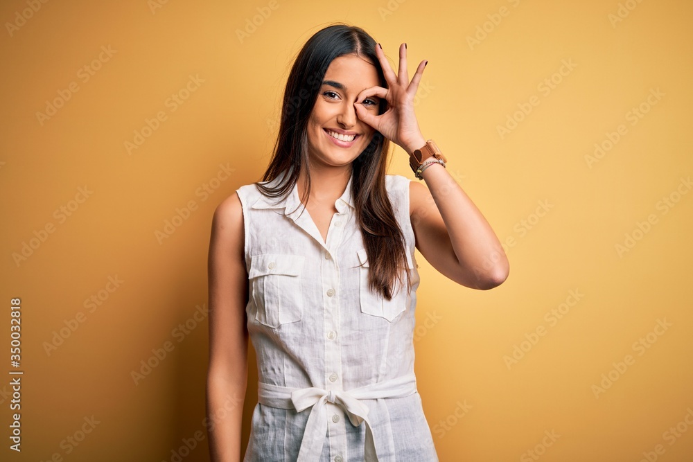 Young beautiful brunette woman on vacation wearing casual dress over yellow background doing ok gesture with hand smiling, eye looking through fingers with happy face. Young beautiful brunette woman on vacation wearing casual dress over yellow background doing ok gesture with hand smiling, eye looking through fingers with happy face.