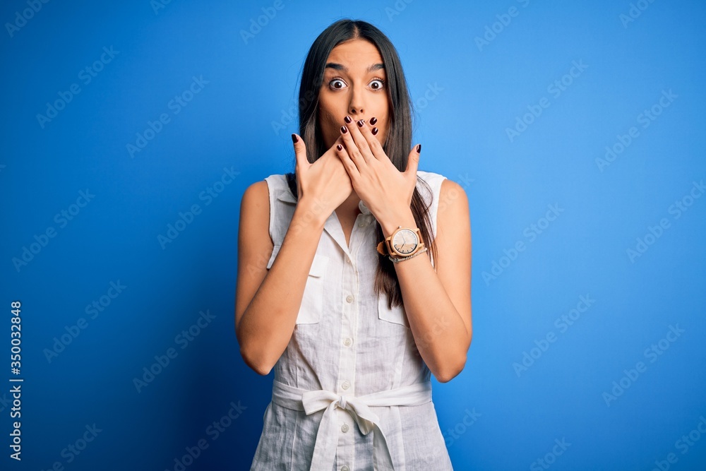 Young beautiful brunette woman wearing casual dress over isolated blue background shocked covering mouth with hands for mistake. Secret concept. Young beautiful brunette woman wearing casual dress over isolated blue background shocked covering mouth with hands for mistake. Secret concept.