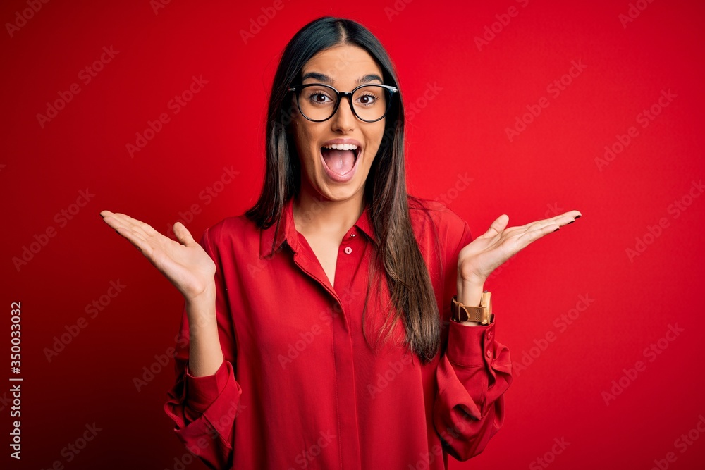 Φωτογραφία Young beautiful brunette woman wearing casual shirt and glasses over red background celebrating crazy and amazed for success with arms raised and open eyes screaming excited