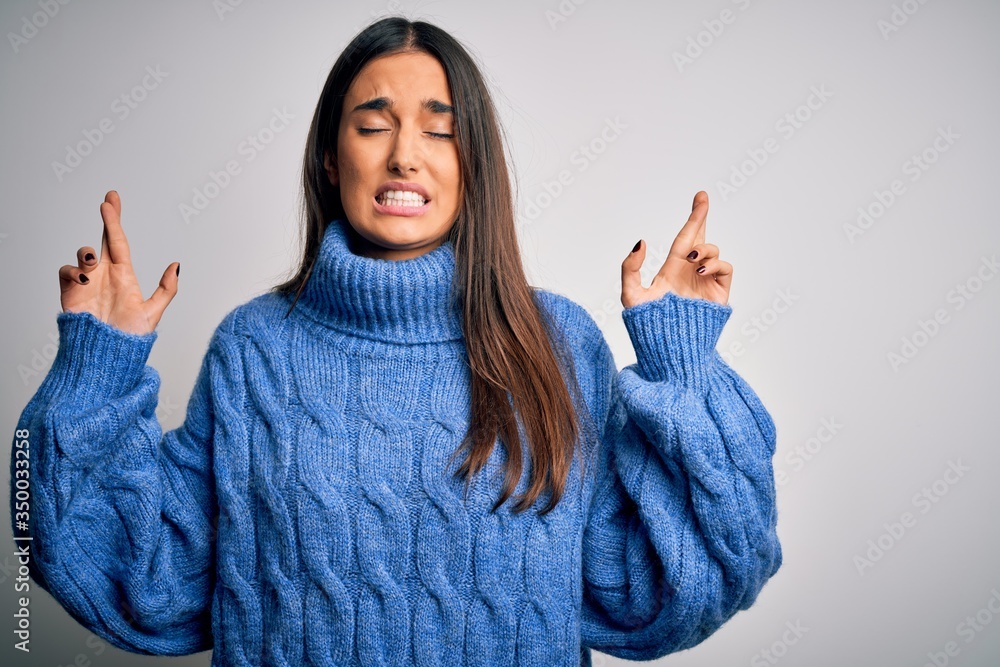 Young beautiful brunette woman wearing casual turtleneck sweater over white background gesturing finger crossed smiling with hope and eyes closed. Luck and superstitious concept. Young beautiful brunette woman wearing casual turtleneck sweater over white background gesturing finger crossed smiling with hope and eyes closed. Luck and superstitious concept.