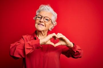 Senior beautiful grey-haired woman wearing casual shirt and glasses over red background smiling in love doing heart symbol shape with hands. Romantic concept.