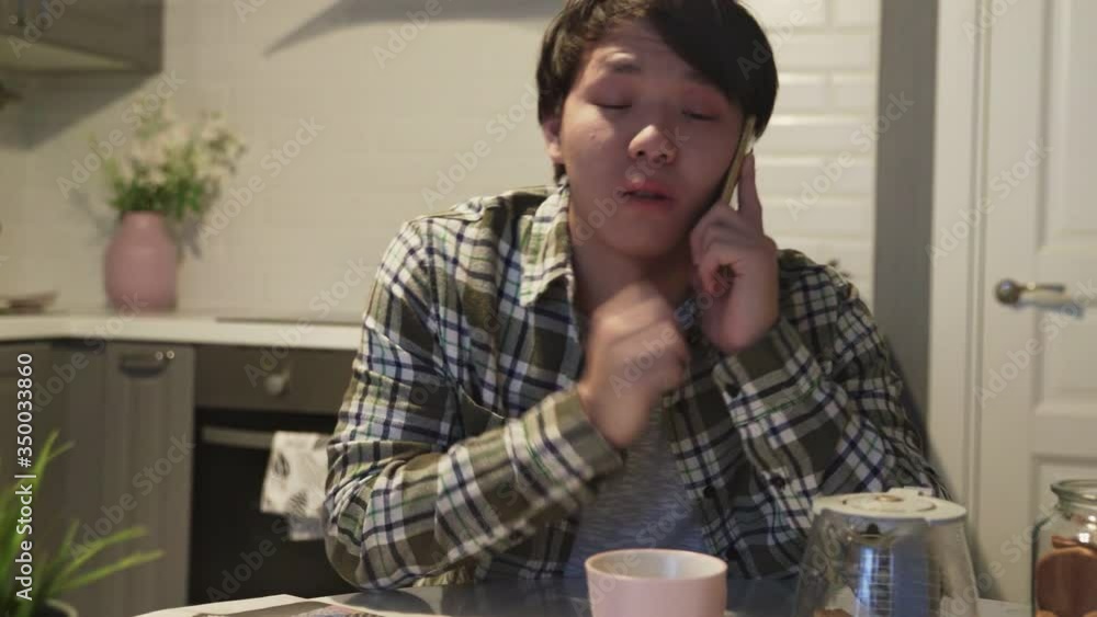 Sleepy asian man talking on mobile phone in kitchen at home Sleepy asian man talking on mobile phone in kitchen at home