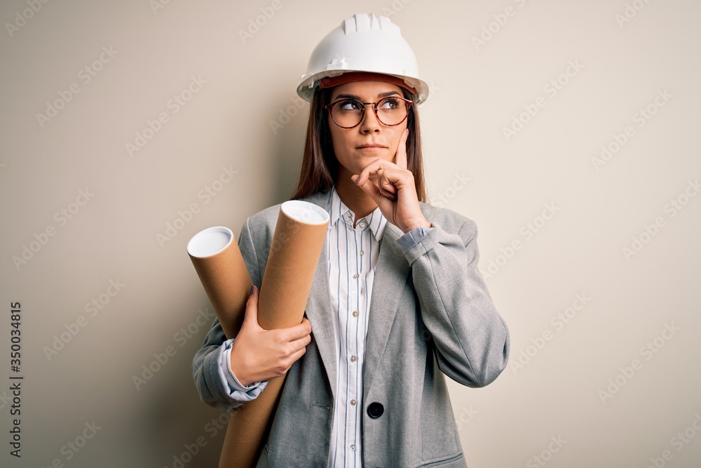 Young beautiful architect woman wearing safety helmet and glasses holding blueprints serious face thinking about question, very confused idea Young beautiful architect woman wearing safety helmet and glasses holding blueprints serious face thinking about question, very confused idea