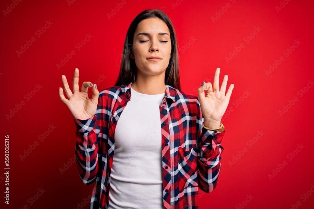 Young beautiful brunette woman wearing casual shirt standing over isolated red background relax and smiling with eyes closed doing meditation gesture with fingers. Yoga concept. Young beautiful brunette woman wearing casual shirt standing over isolated red background relax and smiling with eyes closed doing meditation gesture with fingers. Yoga concept.