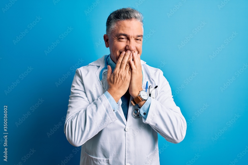 Middle age handsome grey-haired doctor man wearing coat and blue stethoscope laughing and embarrassed giggle covering mouth with hands, gossip and scandal concept Middle age handsome grey-haired doctor man wearing coat and blue stethoscope laughing and embarrassed giggle covering mouth with hands, gossip and scandal concept