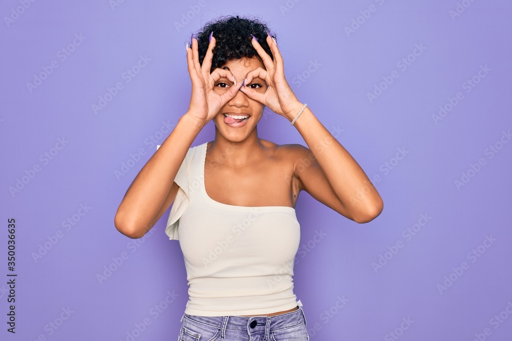 Young beautiful african american afro woman wearing casual t-shirt over purple background doing ok gesture like binoculars sticking tongue out, eyes looking through fingers. Crazy expression. Young beautiful african american afro woman wearing casual t-shirt over purple background doing ok gesture like binoculars sticking tongue out, eyes looking through fingers. Crazy expression.
