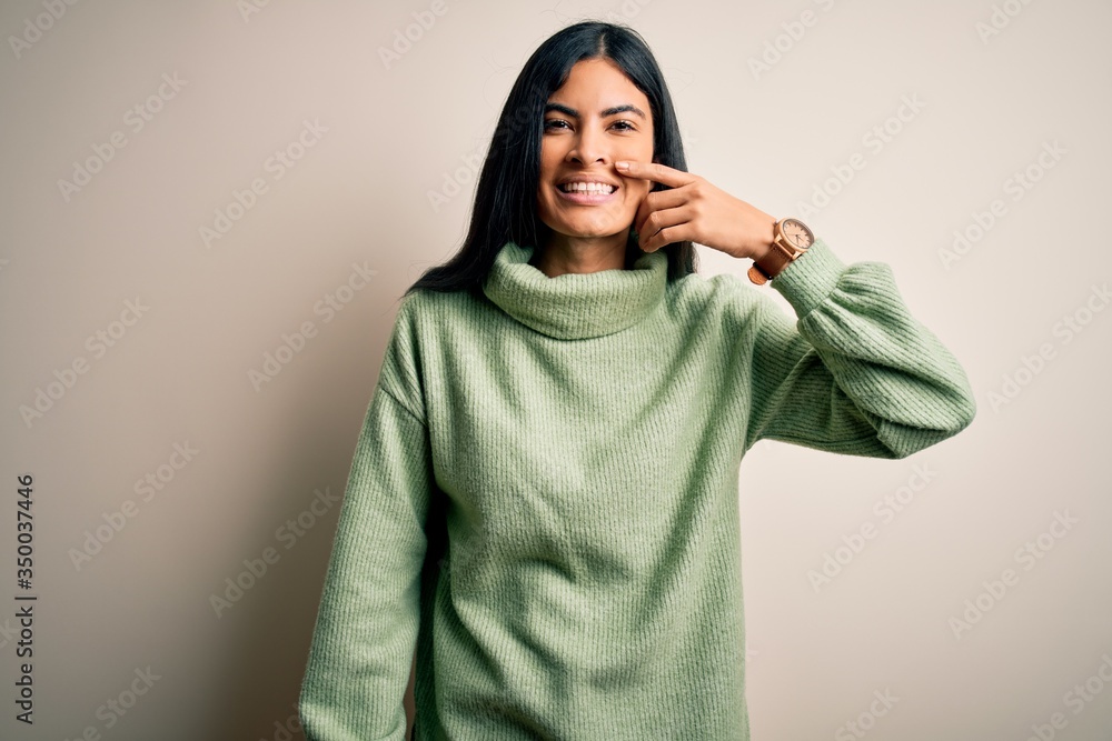 Young beautiful hispanic woman wearing green winter sweater over isolated background Pointing with hand finger to face and nose, smiling cheerful. Beauty concept Young beautiful hispanic woman wearing green winter sweater over isolated background Pointing with hand finger to face and nose, smiling cheerful. Beauty concept