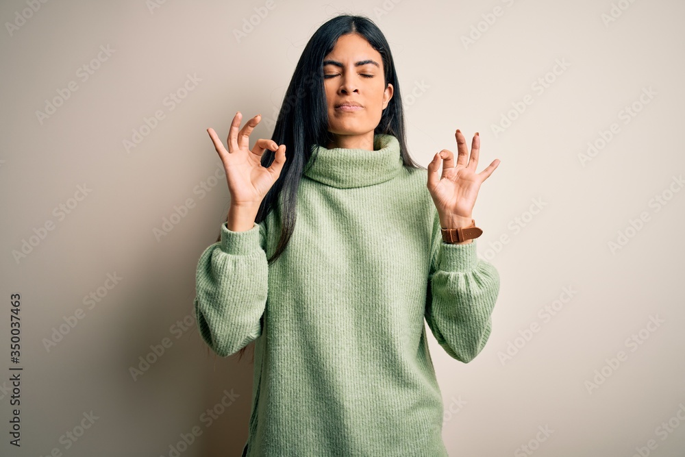Young beautiful hispanic woman wearing green winter sweater over isolated background relaxed and smiling with eyes closed doing meditation gesture with fingers. Yoga concept. Young beautiful hispanic woman wearing green winter sweater over isolated background relaxed and smiling with eyes closed doing meditation gesture with fingers. Yoga concept.