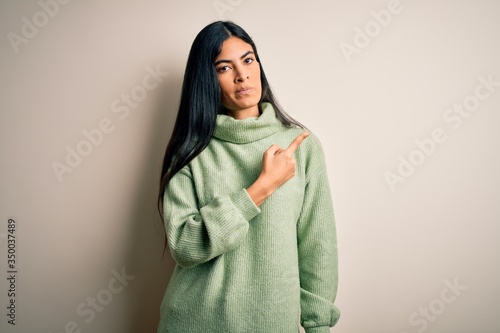 Young beautiful hispanic woman wearing green winter sweater over isolated background Pointing with hand finger to the side showing advertisement, serious and calm face © Krakenimages.com