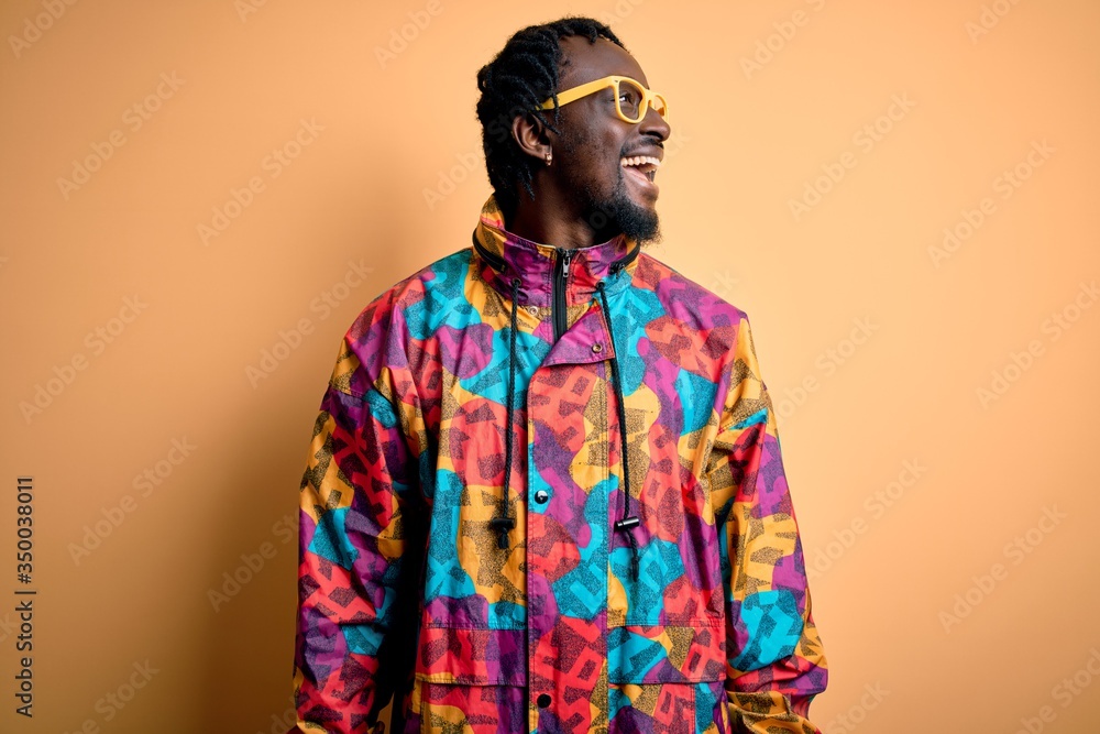 Handsome african american man wearing colorful coat and glasses over yellow background looking away to side with smile on face, natural expression. Laughing confident.