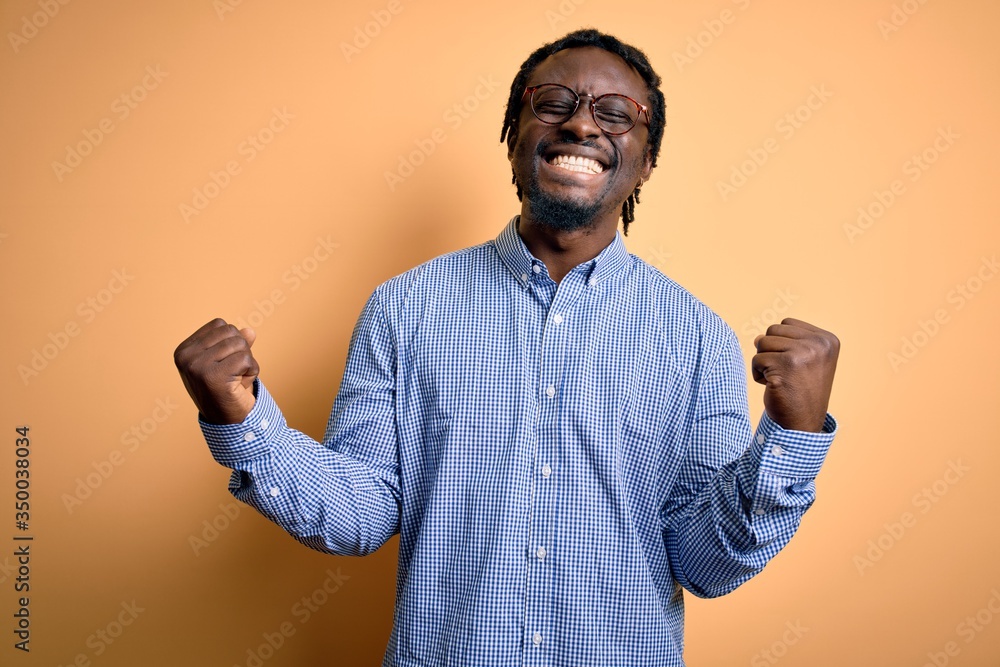 Young handsome african american man wearing shirt and glasses over yellow background very happy and excited doing winner gesture with arms raised, smiling and screaming for success. Celebration Young handsome african american man wearing shirt and glasses over yellow background very happy and excited doing winner gesture with arms raised, smiling and screaming for success. Celebration
