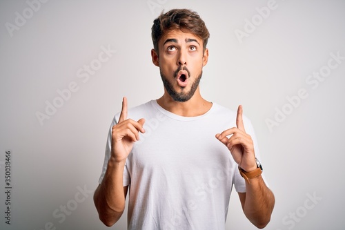 Young handsome man with beard wearing casual t-shirt standing over white background amazed and surprised looking up and pointing with fingers and raised arms.