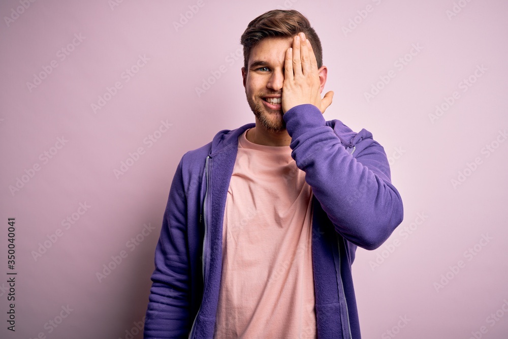 Young blond man with beard and blue eyes wearing purple sweatshirt over pink background covering one eye with hand, confident smile on face and surprise emotion.