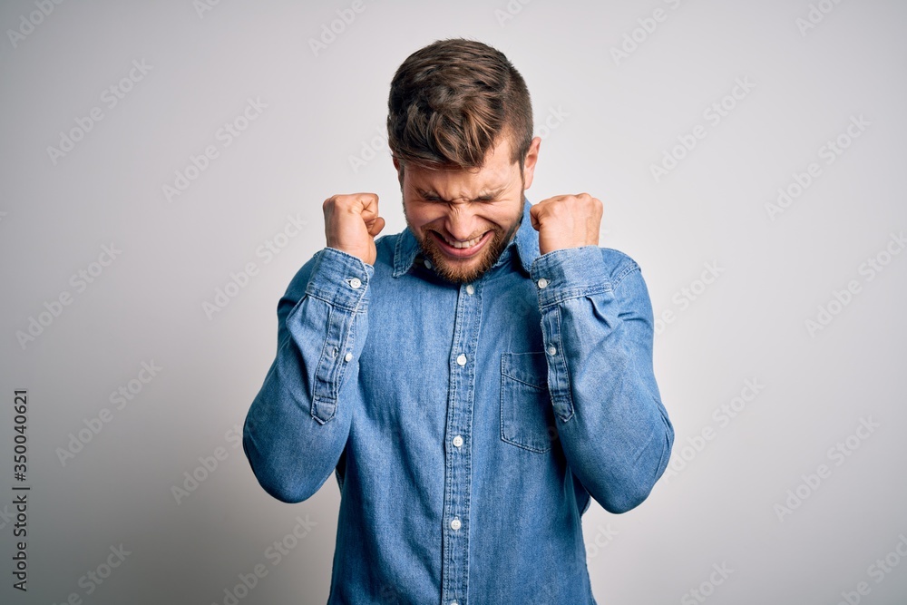 Young handsome blond man with beard and blue eyes wearing casual denim shirt excited for success with arms raised and eyes closed celebrating victory smiling. Winner concept. Young handsome blond man with beard and blue eyes wearing casual denim shirt excited for success with arms raised and eyes closed celebrating victory smiling. Winner concept.