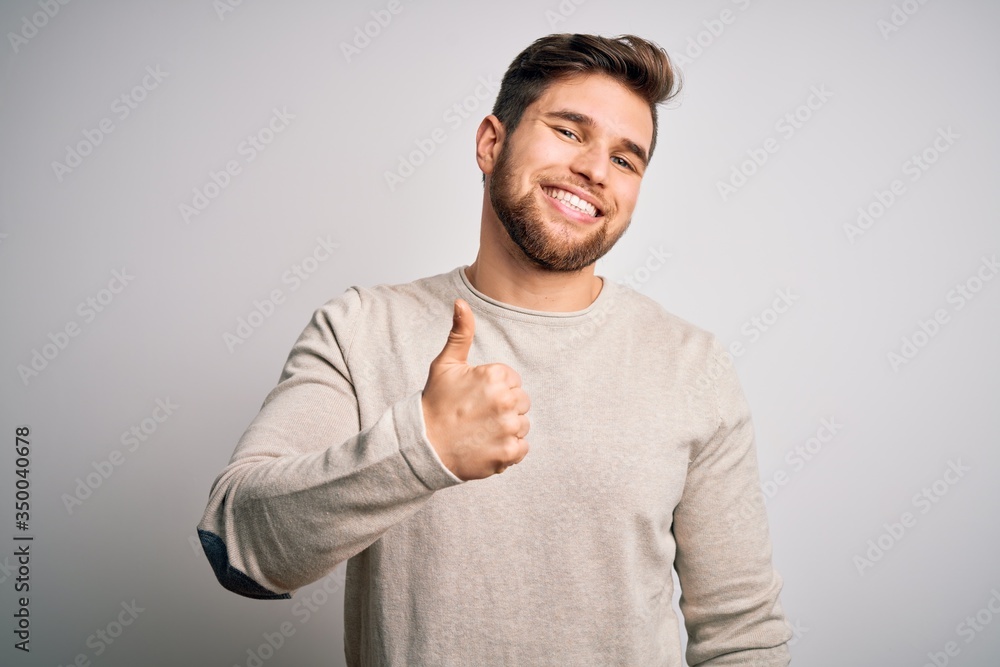 Young handsome blond man with beard and blue eyes wearing casual sweater doing happy thumbs up gesture with hand. Approving expression looking at the camera showing success. Young handsome blond man with beard and blue eyes wearing casual sweater doing happy thumbs up gesture with hand. Approving expression looking at the camera showing success.