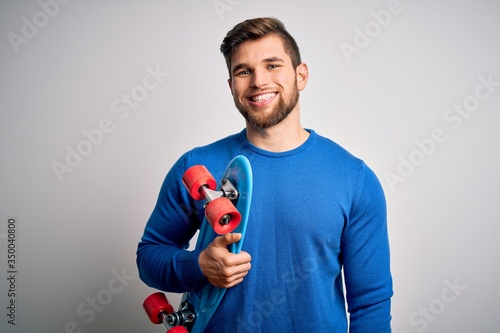 Young blond skeater man with beard and blue eyes holding skate over white background with a happy face standing and smiling with a confident smile showing teeth
