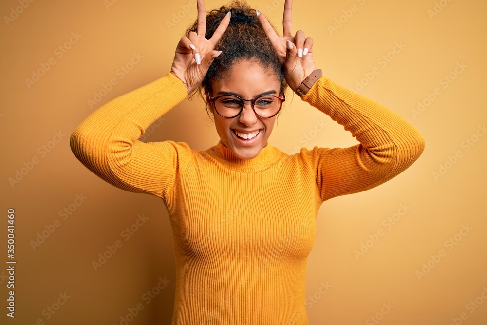 Young beautiful african american girl wearing sweater and glasses over yellow background Posing funny and crazy with fingers on head as bunny ears, smiling cheerful Young beautiful african american girl wearing sweater and glasses over yellow background Posing funny and crazy with fingers on head as bunny ears, smiling cheerful