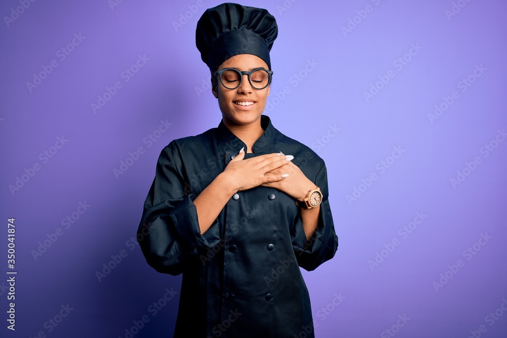 Young african american chef girl wearing cooker uniform and hat over purple background smiling with hands on chest with closed eyes and grateful gesture on face. Health concept. Young african american chef girl wearing cooker uniform and hat over purple background smiling with hands on chest with closed eyes and grateful gesture on face. Health concept.