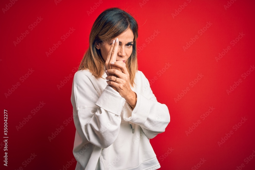 Middle age beautiful sportswoman wearing casual sweatshirt over isolated red background Holding symbolic gun with hand gesture, playing killing shooting weapons, angry face