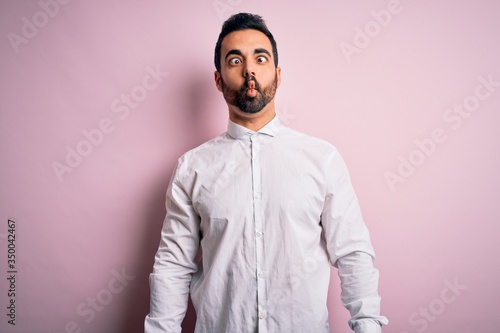 Young handsome man with beard wearing casual shirt standing over pink background making fish face with lips, crazy and comical gesture. Funny expression.