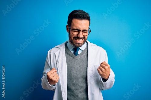 Young handsome doctor man with beard wearing coat and glasses over blue background very happy and excited doing winner gesture with arms raised, smiling and screaming for success. Celebration concept.