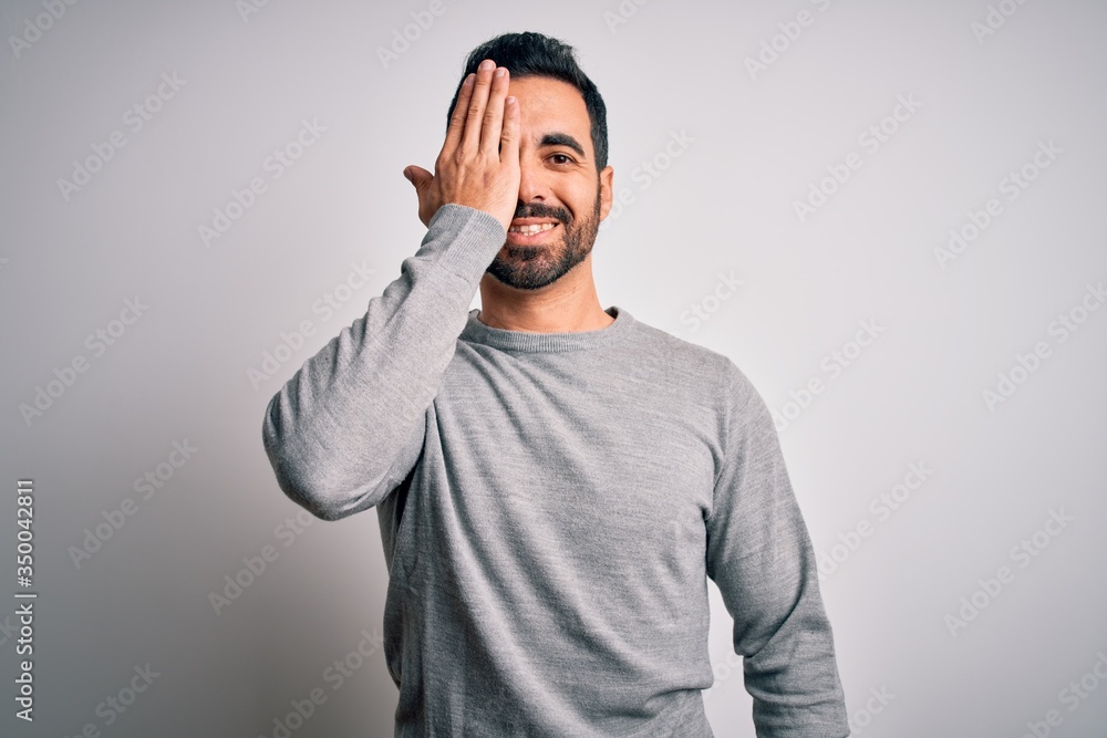 Young handsome man with beard wearing casual sweater standing over white background covering one eye with hand, confident smile on face and surprise emotion. Young handsome man with beard wearing casual sweater standing over white background covering one eye with hand, confident smile on face and surprise emotion.