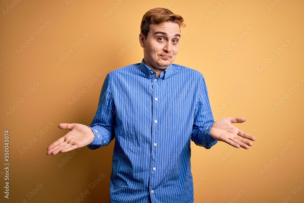 Young handsome redhead man wearing casual striped shirt over isolated yellow background clueless and confused expression with arms and hands raised. Doubt concept. Young handsome redhead man wearing casual striped shirt over isolated yellow background clueless and confused expression with arms and hands raised. Doubt concept.