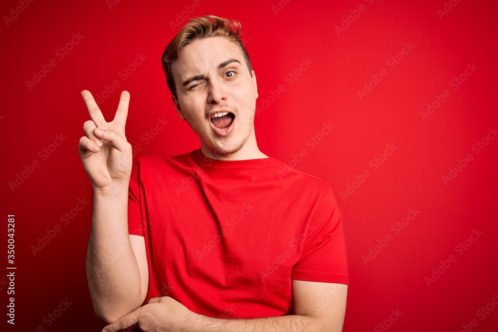 Naklejka premium Young handsome redhead man wearing casual t-shirt over isolated red background smiling with happy face winking at the camera doing victory sign. Number two.